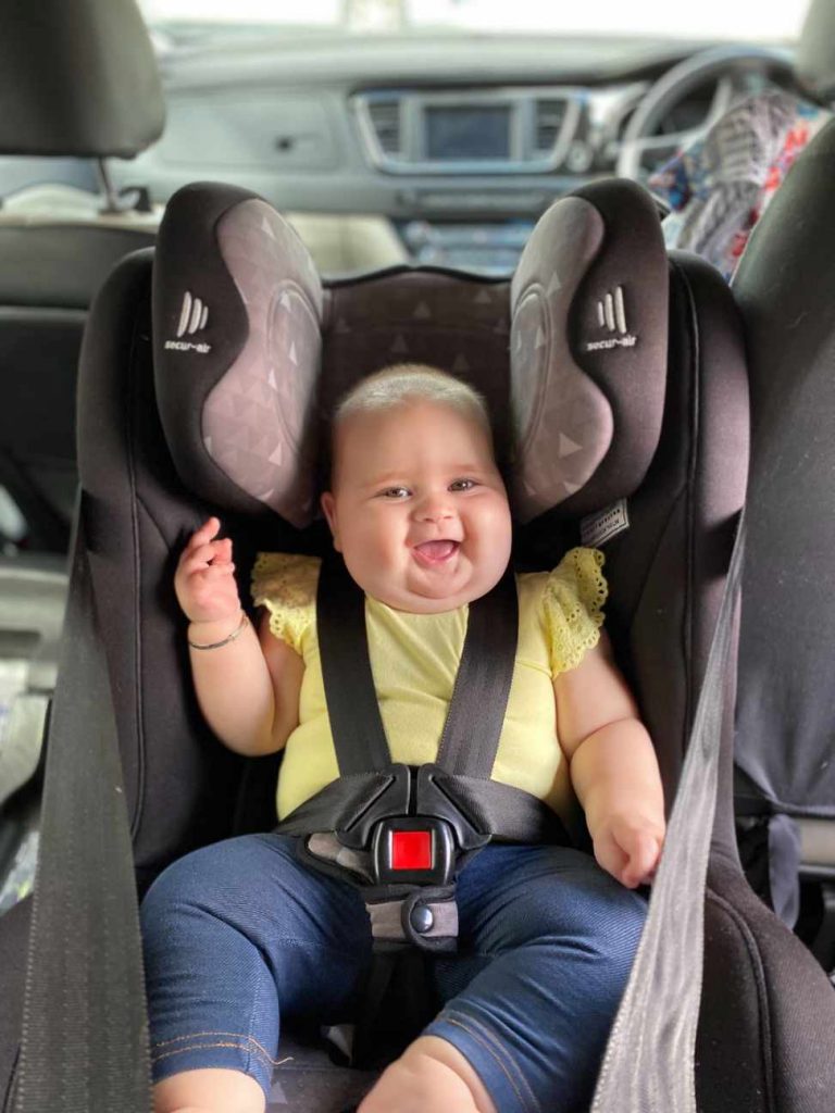 Smiling baby in a rear facing car seat wearing a yellow shirt and blue jeans